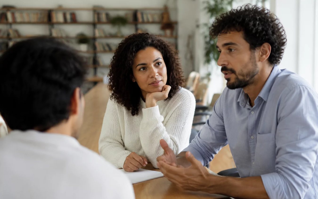 Três participantes conversam ao redor de uma mesa comprida numa sala clara de janelas altas